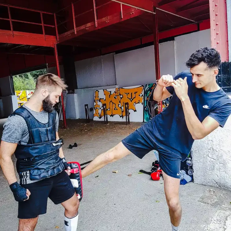 Entraînement de boxe pieds-poings en extérieur avec un pratiquant donnant un coup de pied sur une cible tenue par le coach.