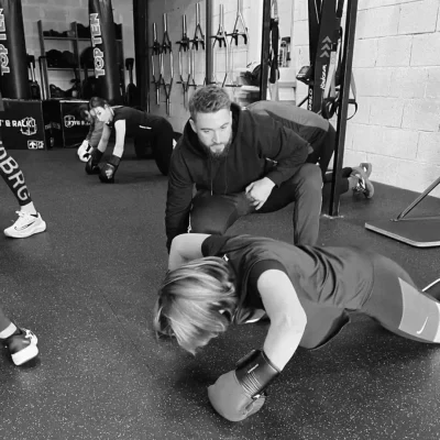Coach sportif guidant une cliente lors d’un exercice de renforcement au sol, gants de boxe aux mains, dans une salle d’entraînement.